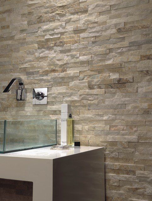 Badezimmer mit Steinfliesenwand, modernem Wasserhahn und Glaswaschbecken, weißer Arbeitsplatte, Toilettenartikeln.