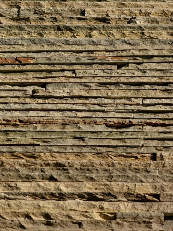 Nahaufnahme einer verwitterten Steinmauer mit horizontalen Schichten in Beige- und Grautönen.