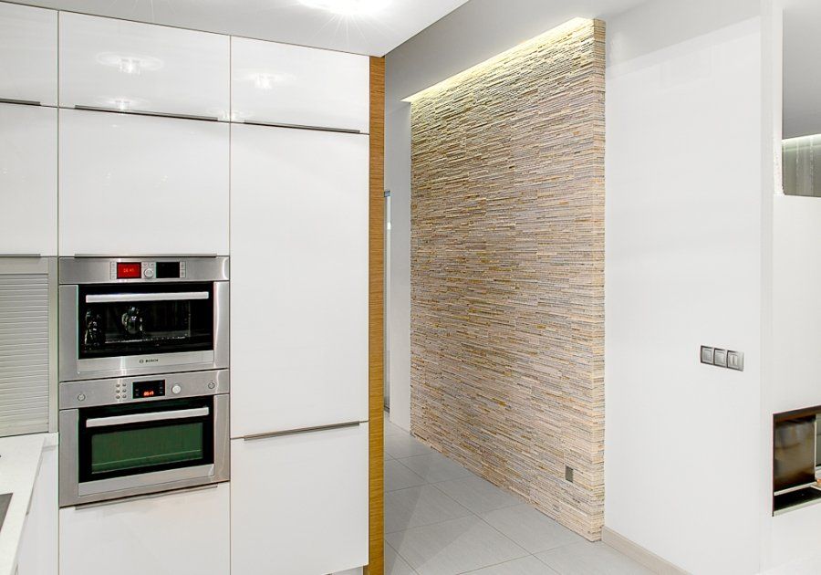 Moderne weiße Küche mit eingebauten Edelstahlöfen und Akzentwand aus strukturiertem Stein.