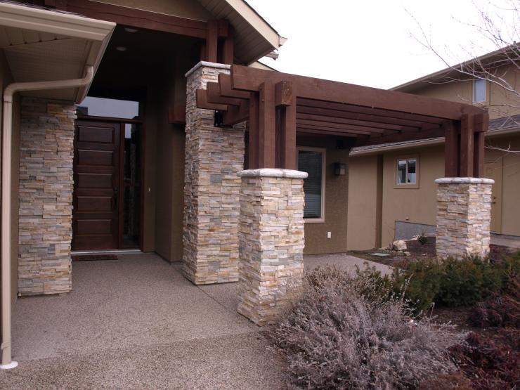 Steinsäulen und Pergola über der Veranda. Dunkelbraune Balken und Tür. Beige Fassadenverkleidung, Betonweg.