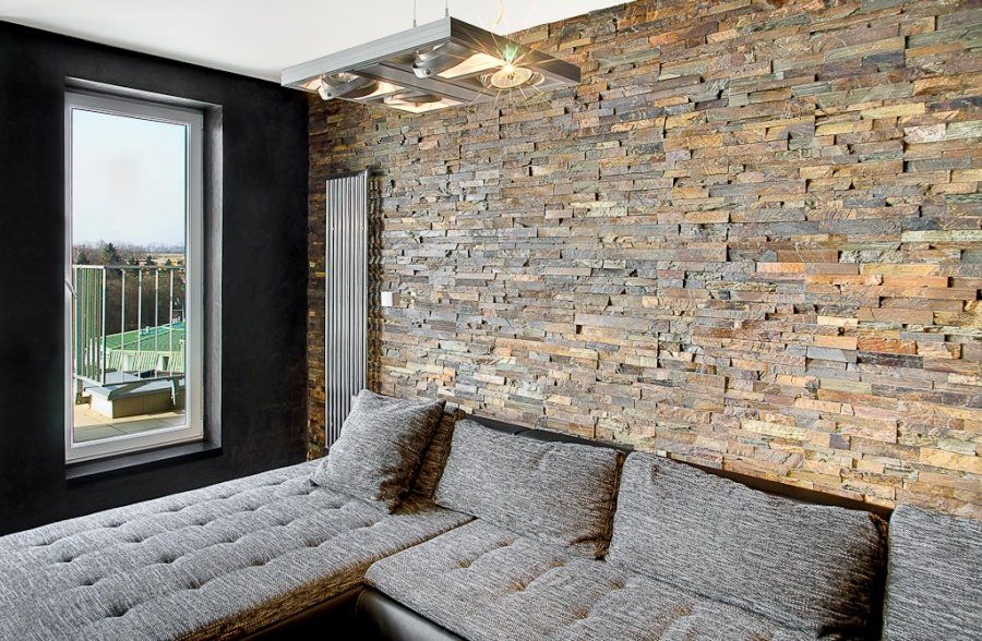 Wohnzimmer mit Steinwand als Akzent, grauem Ecksofa und Fenster mit Blick auf einen Balkon.