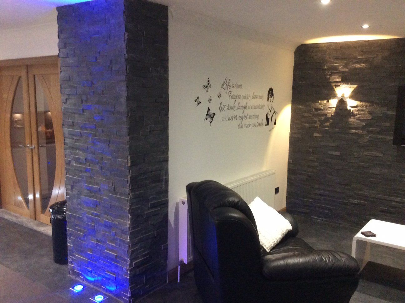 Wohnzimmer mit Steinsäule und Akzentwand. Ein schwarzer Ledersessel steht einem weißen Tisch gegenüber.