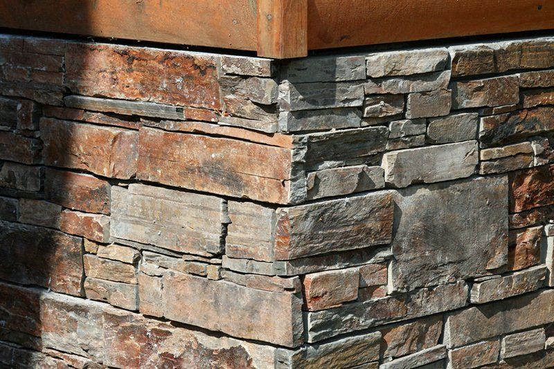 Ecke einer Steinmauer aus rechteckigen Steinen in verschiedenen Braun-, Grau- und Beigetönen, unter einem braunen Holzbalken.