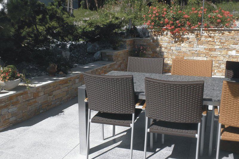 Essgruppe für die Terrasse mit geflochtenen Stühlen und einer Steinmauer in sonniger Außenlage.