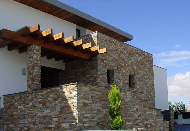 Modernes Hausäußeres mit Steinfassade, Holzpergola und kleinem immergrünen Baum.