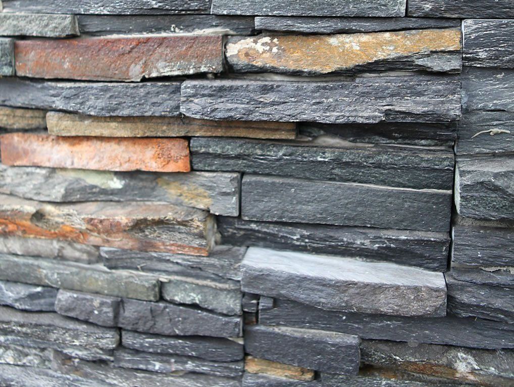 Nahaufnahme einer Steinmauer aus rechteckigen, mehrfarbigen Schieferplatten in Grau-, Braun- und Orangetönen.