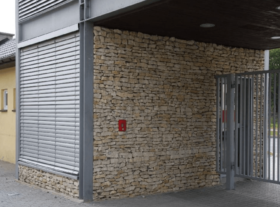 Die Fassade des Gebäudes ist von Steinmauern umgeben und mit einem geschlossenen Metalltor versehen. Links befindet sich ein Fenster mit geschlossenen Jalousien.