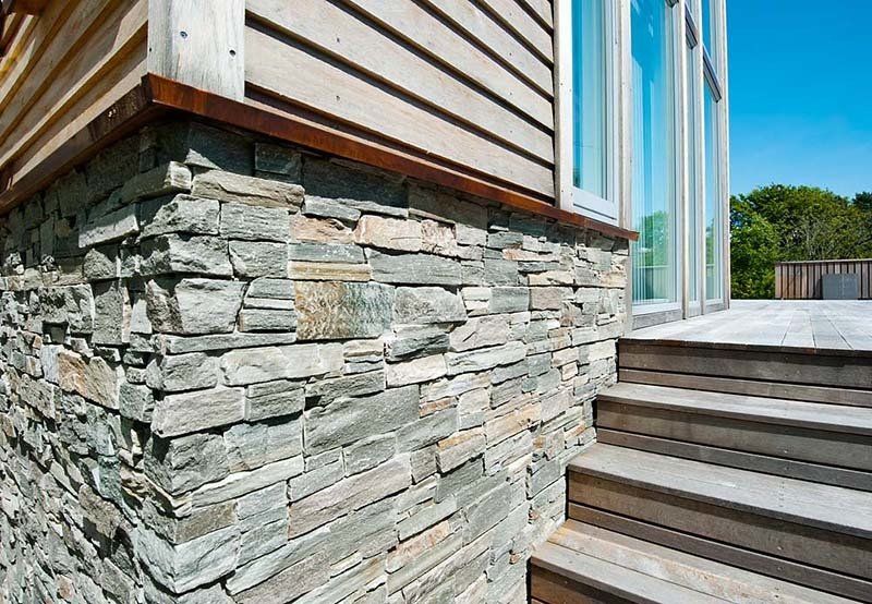 Eine Hausecke ist mit einer Steinverblendung und Holzverkleidung versehen, dazu kommen Holztreppen und Holzfenster.