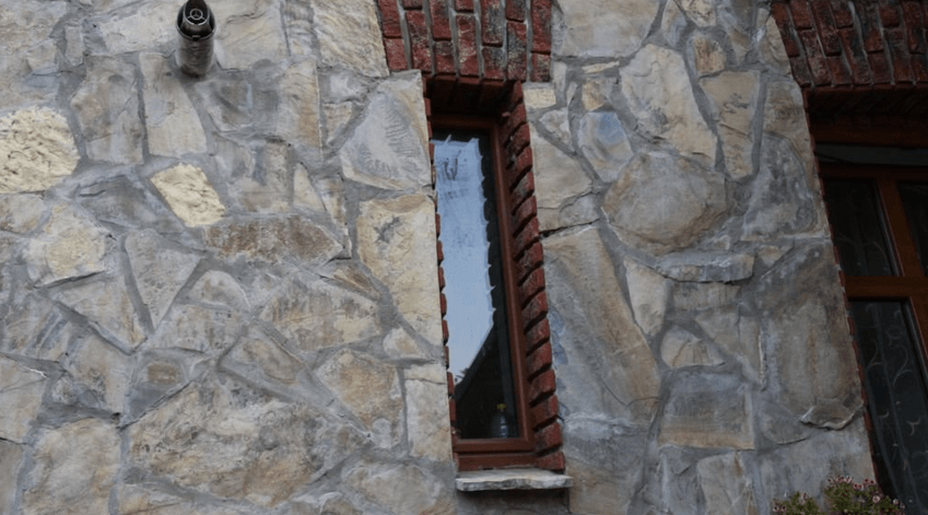 Außenfassade aus Stein mit hohem, schmalem Fenster und braunem Rahmen. Oben roter Backstein.