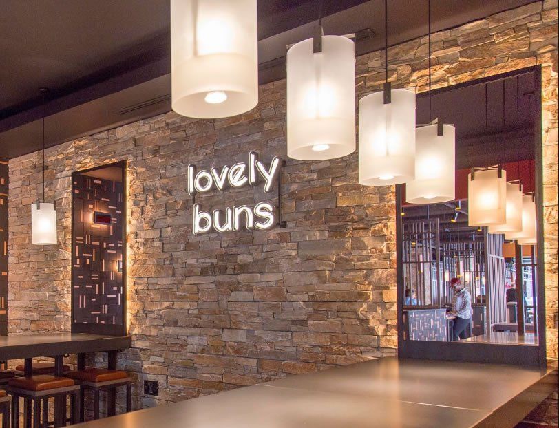 Restaurantinterieur mit Steinwand, „Lovely Buns“-Schild und Pendelleuchten. Bar mit Barhockern sichtbar.