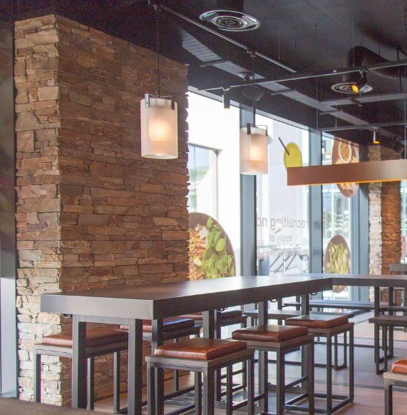 Restaurantinterieur mit Steinwand, langem Tisch, Hockern und Pendelleuchten.