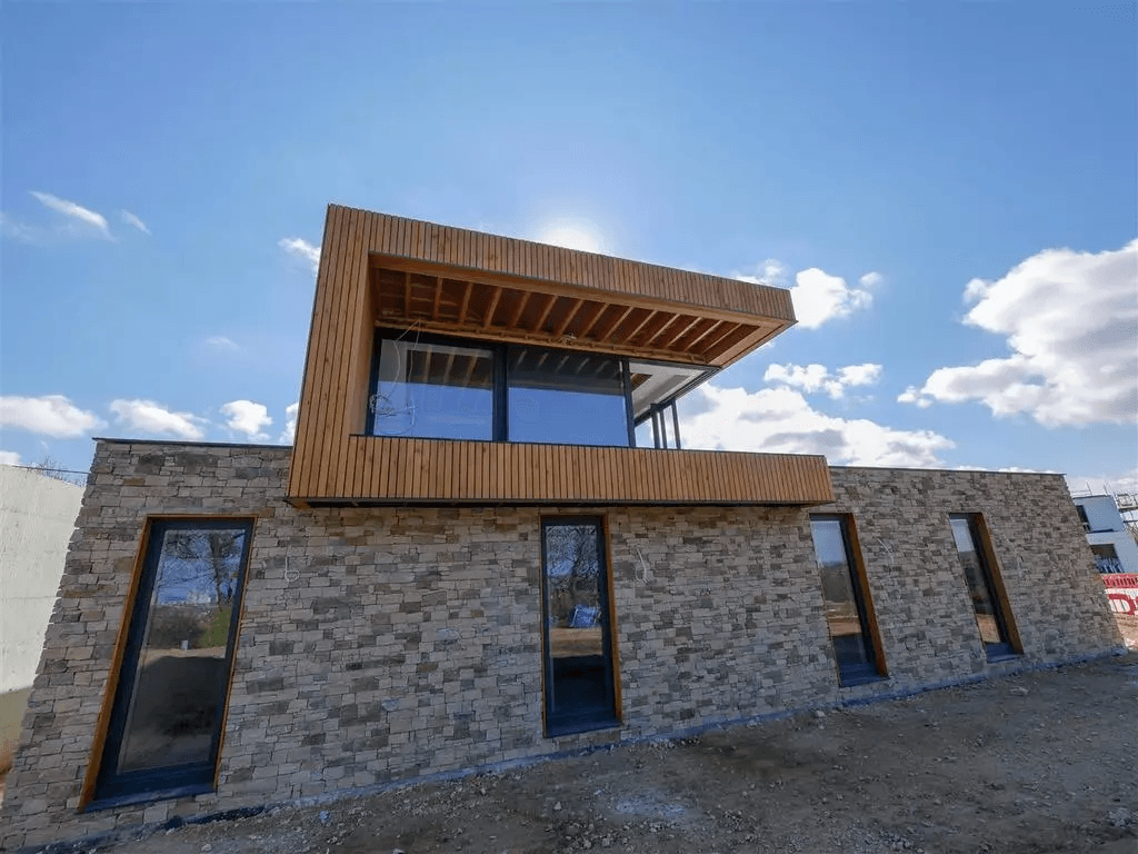 Modernes Haus mit Steinfassade, Holzelementen und großen Fenstern unter strahlend blauem Himmel.