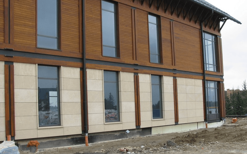 Gebäudeaußenansicht mit brauner Holz- und beiger Steinfassade, hohen rechteckigen Fenstern und einem bewölkten Himmel.