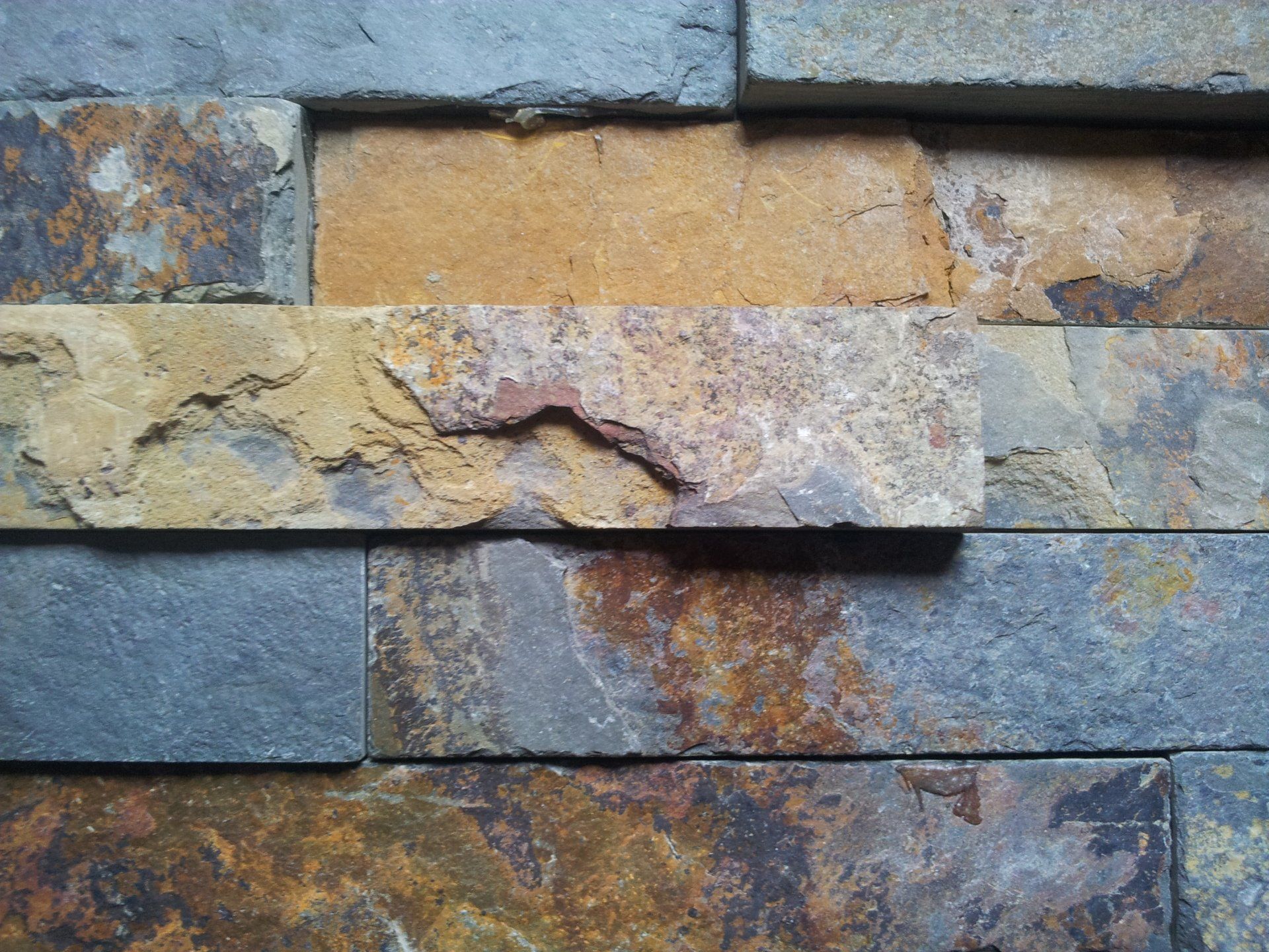 Nahaufnahme einer Wand aus rechteckigen, mehrfarbigen Steinfliesen; Farbtöne von Gold, Braun und Blau.