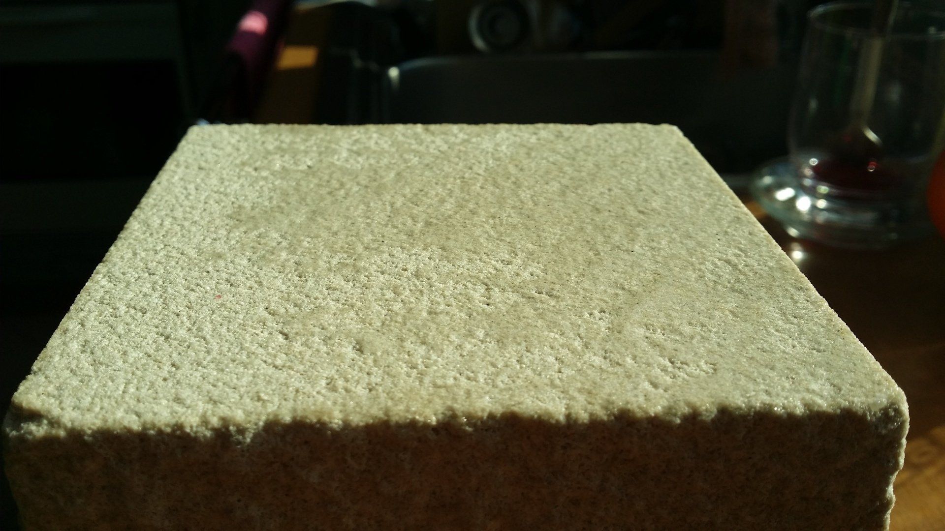 Ein quadratischer, hellbrauner Ziegelstein auf einem Tisch, vom Sonnenlicht beschienen.