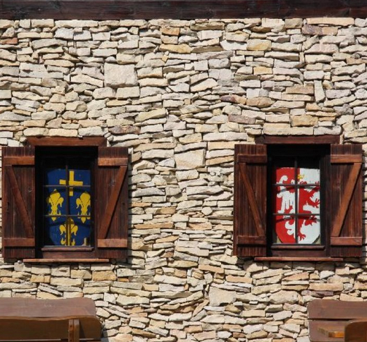 Zwei Fenster mit Holzläden, hinter denen sich Wappen auf einer Steinmauer befinden. Das eine ist blau-golden, das andere rot-weiß.