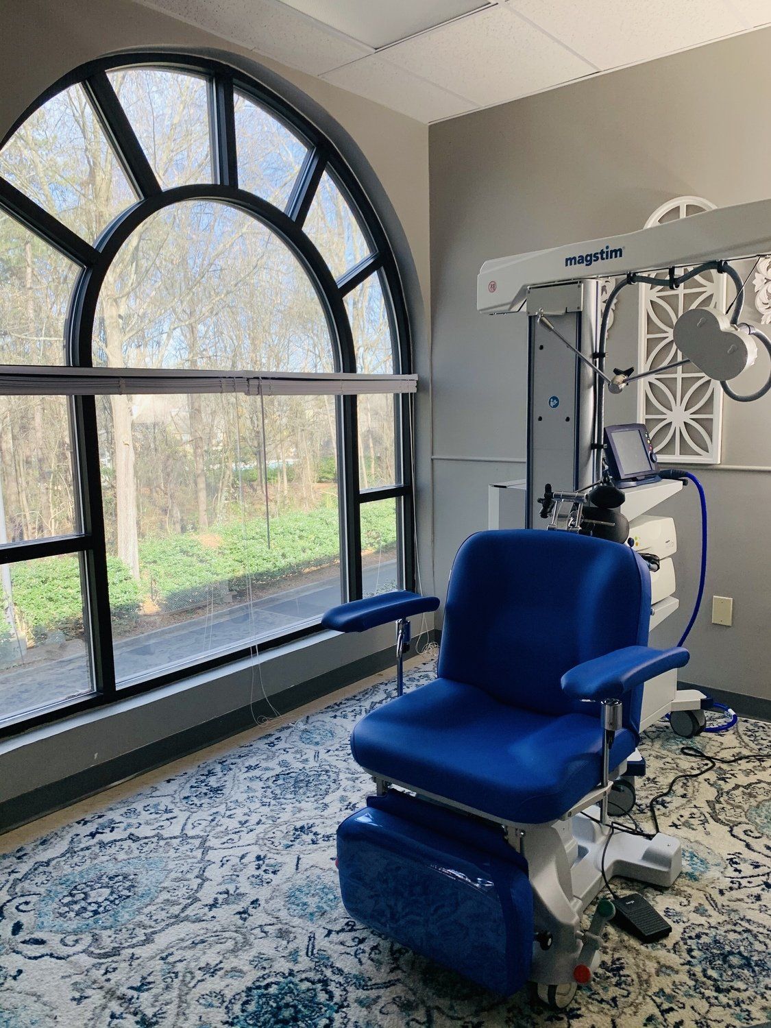 Blue medical chair near a large arched window with a view of trees. The room has grey walls and patterned flooring.