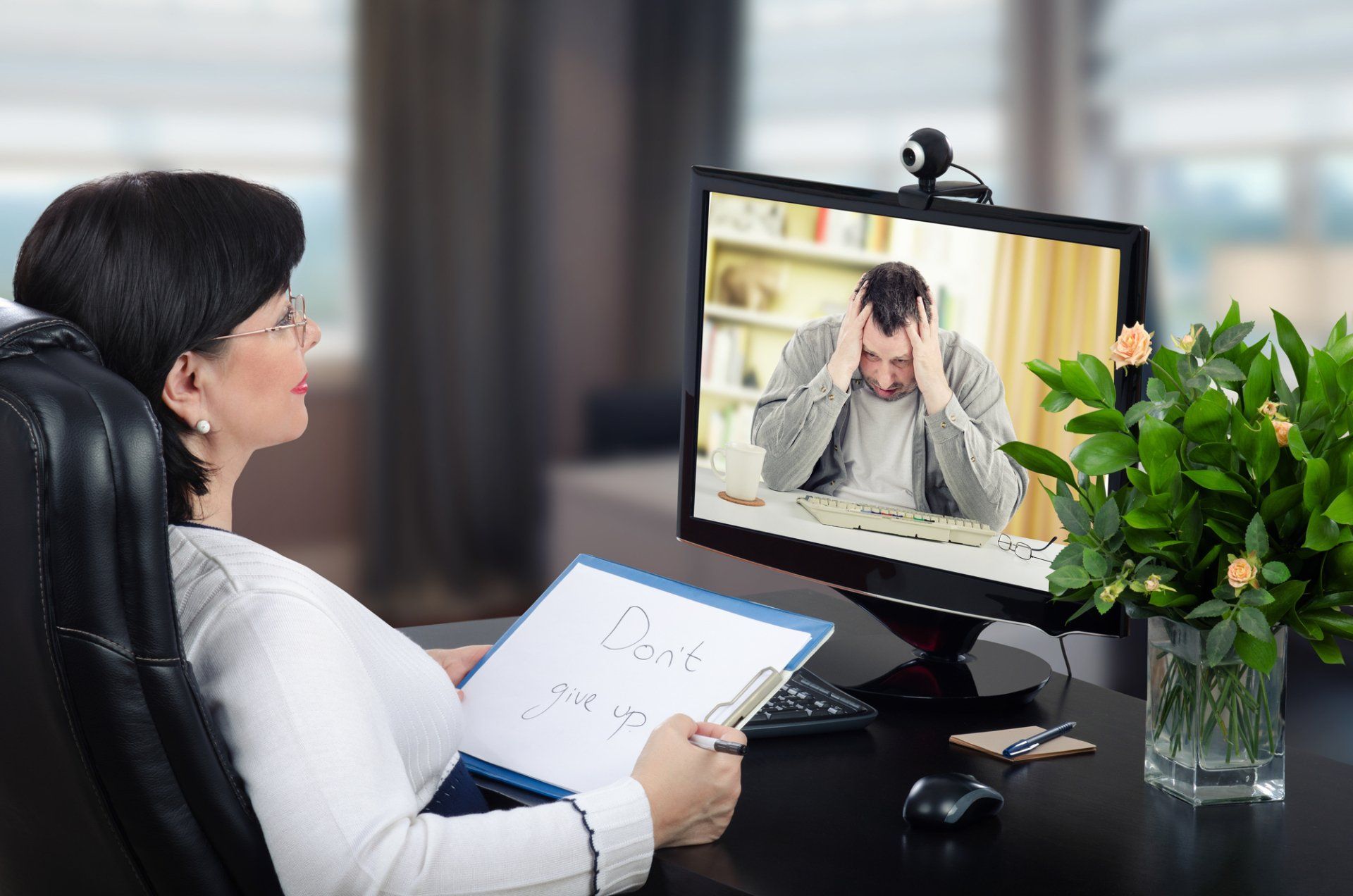 Woman counseling a person via video call; woman holds a notepad, man on screen appears stressed.
