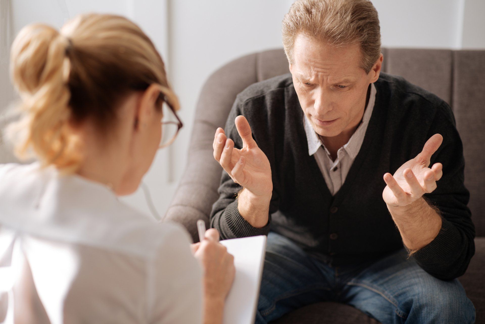 A person gestures while talking to a person holding a notepad, possibly in a therapy setting.