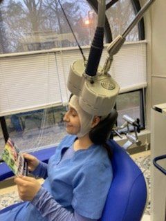 Woman in blue scrubs sits in a chair with a device on her head, possibly receiving medical treatment, in a clinic.