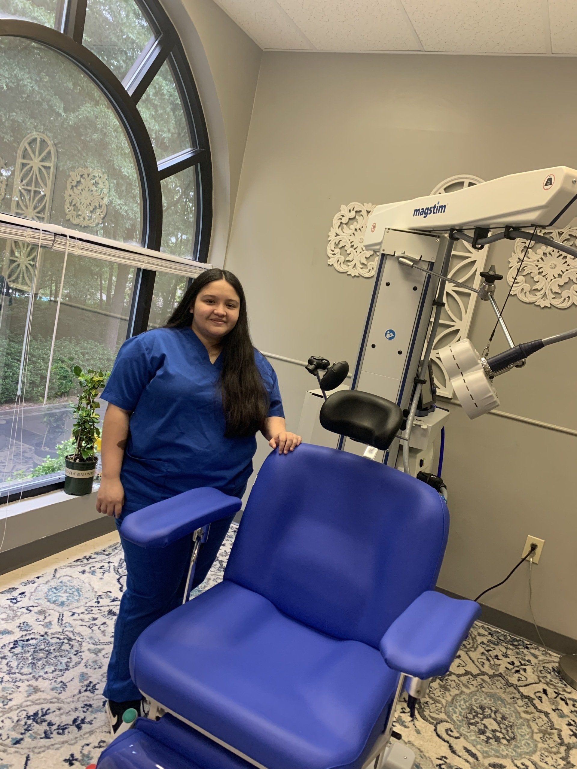 Woman in blue scrubs stands near a blue medical chair and equipment in a room with a window.