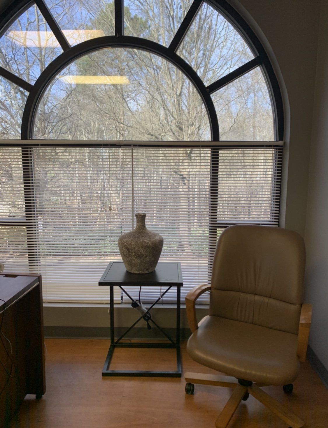 Office interior with arched window, chair, small table with vase, and outdoor view.
