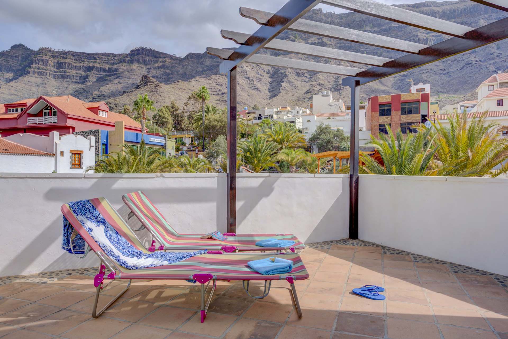 Zwei Liegestühle stehen unter einer Pergola auf einer Terrasse mit Bergen im Hintergrund.