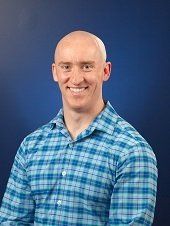 Bald man smiling in a blue plaid shirt against a blue background.