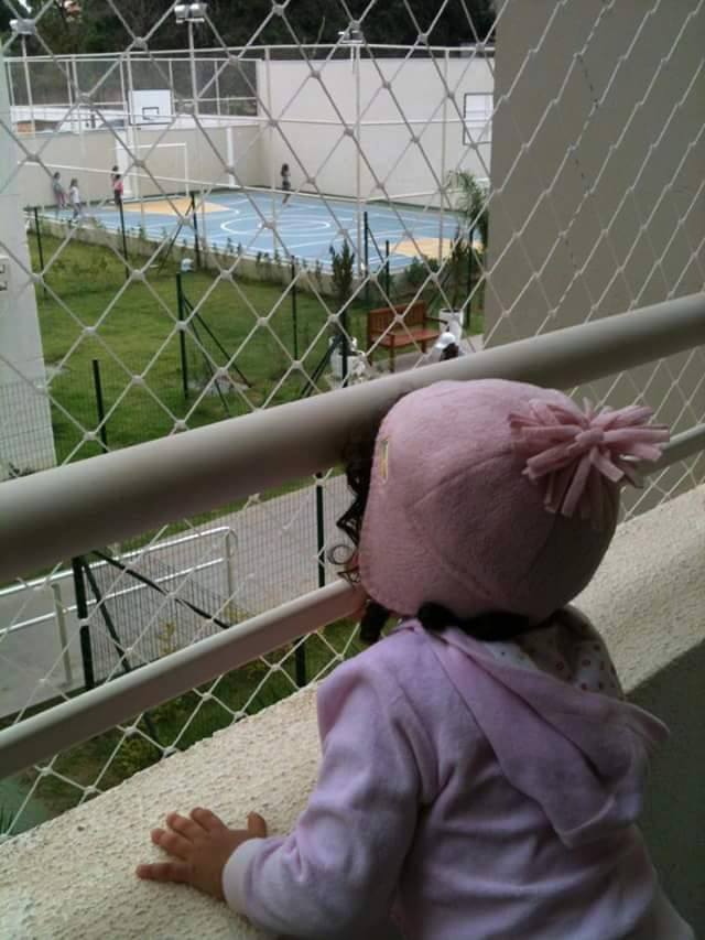 Criança com chapéu rosa olhando através de uma rede para uma piscina e um pátio gramado.
