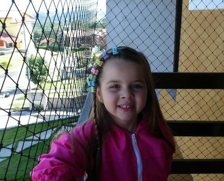 Criança sorridente com jaqueta rosa em pé ao lado de uma cerca de rede em um parquinho infantil.