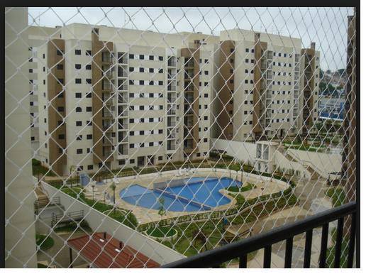 Pátio de um complexo de apartamentos com piscina azul, visto através de uma cerca de arame.