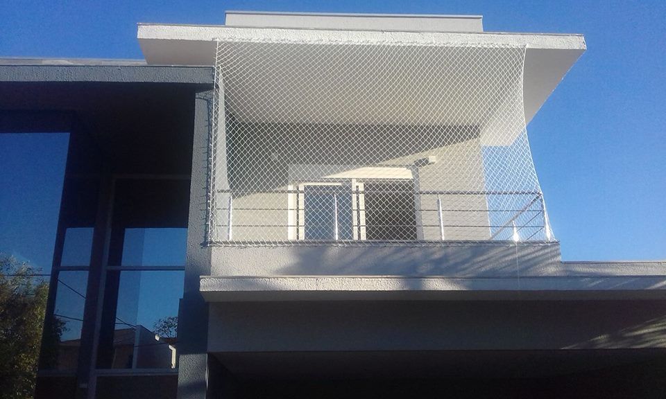 Fachada moderna de edifício branco com varanda, tela metálica e céu azul ao fundo.