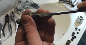 A person uses a screwdriver on a faucet component, near a sink with tools and parts.
