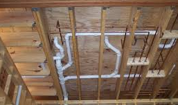 Under-floor view of exposed plumbing pipes: white PVC waste pipes, copper supply pipes, wood beams and subfloor.