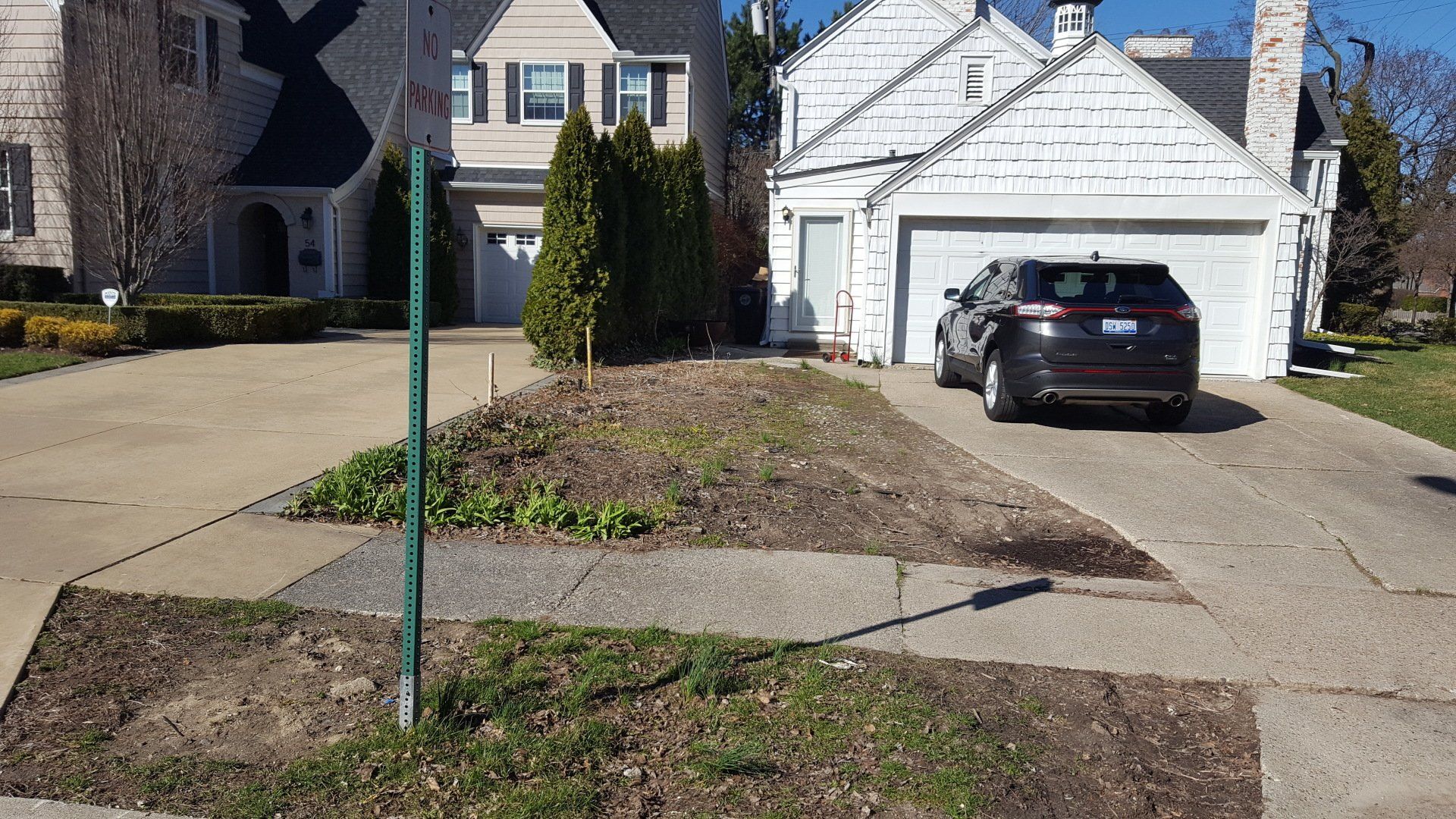 A residential driveway and yard with mulch beds and evergreen bushes, next to a garage with a parked dark SUV.