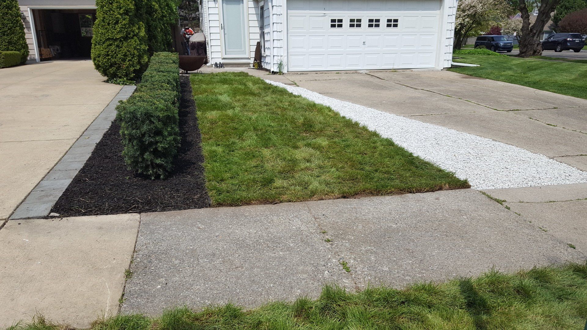 A residential front yard with a mulch garden bed, a patch of green grass, and a decorative white rock strip near a driveway.