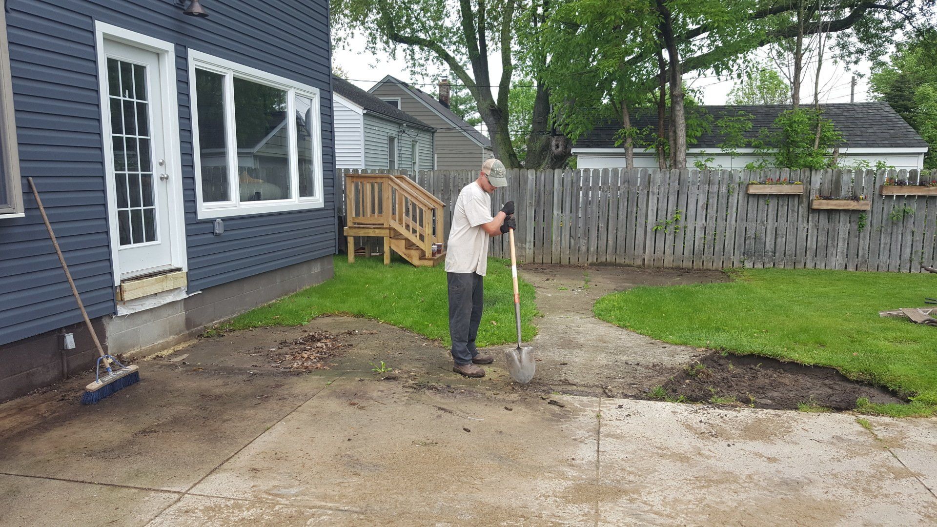A person uses a shovel to clear dirt from a concrete patio area in a grassy backyard next to a dark blue house.