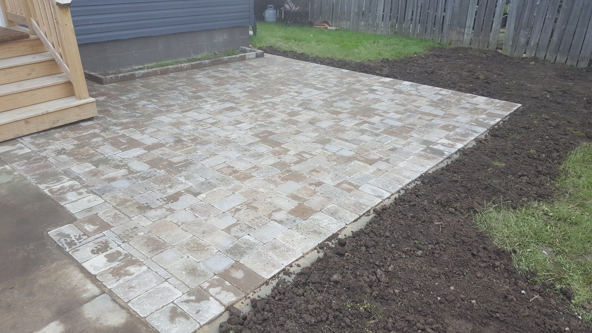 A light-colored stone patio sits adjacent to a wooden staircase and a home exterior, bordered by a patch of dark soil.
