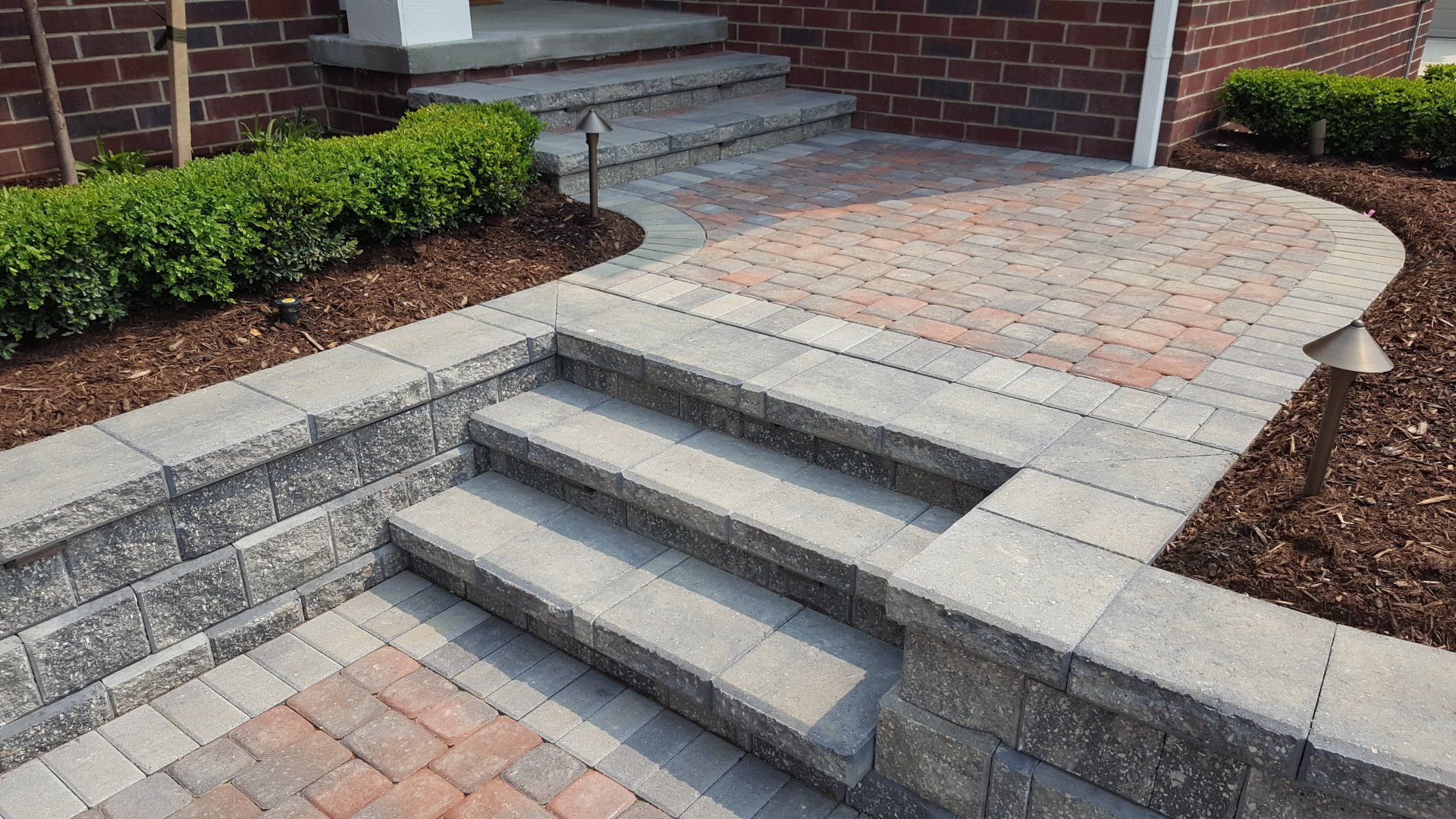 A brick house entrance with a stone paver walkway, two sets of concrete steps, and landscaping with mulch and shrubs.