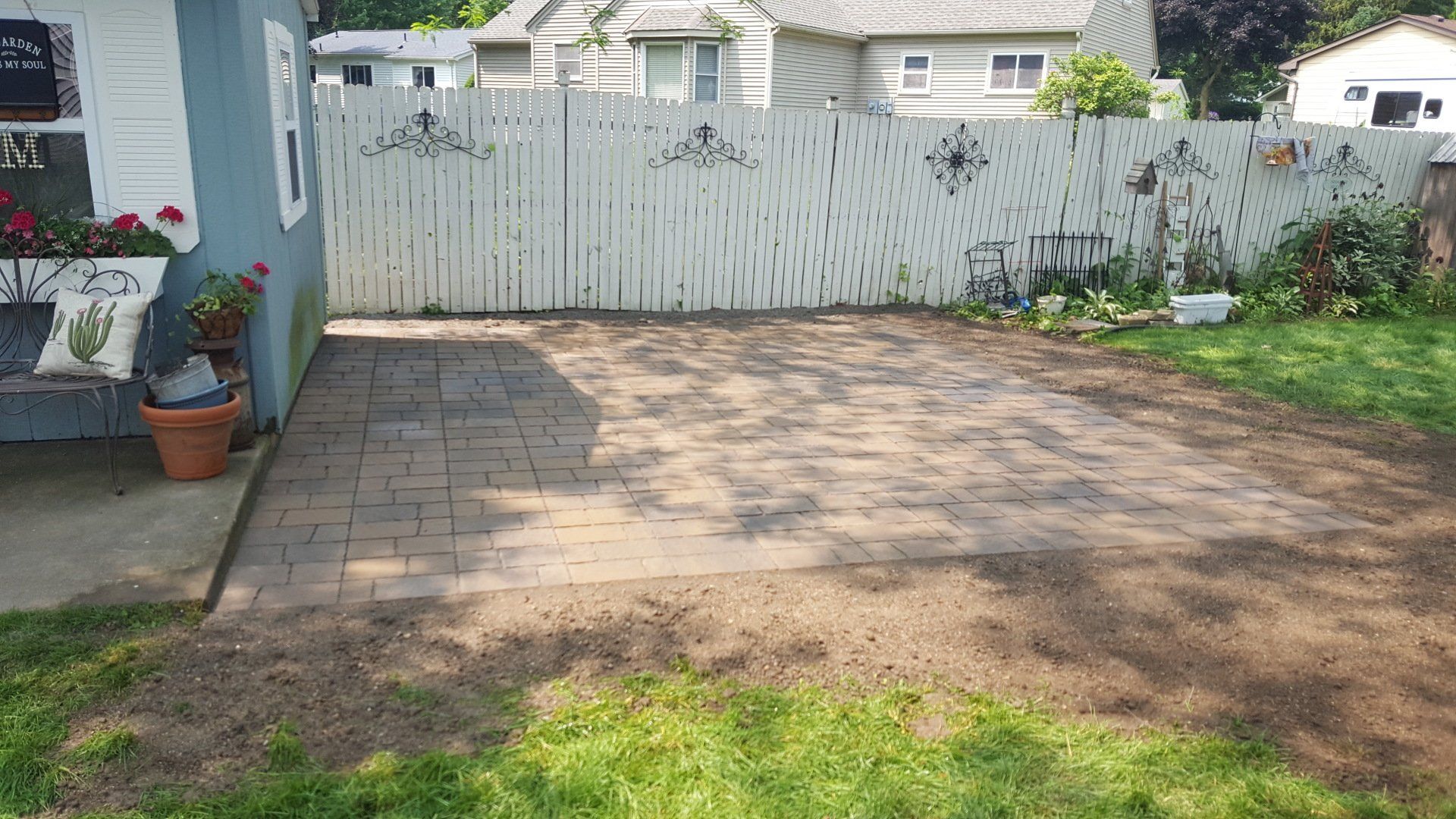 A backyard patio made of rectangular stone pavers, bordered by a white picket fence, a small shed, and dirt patches.