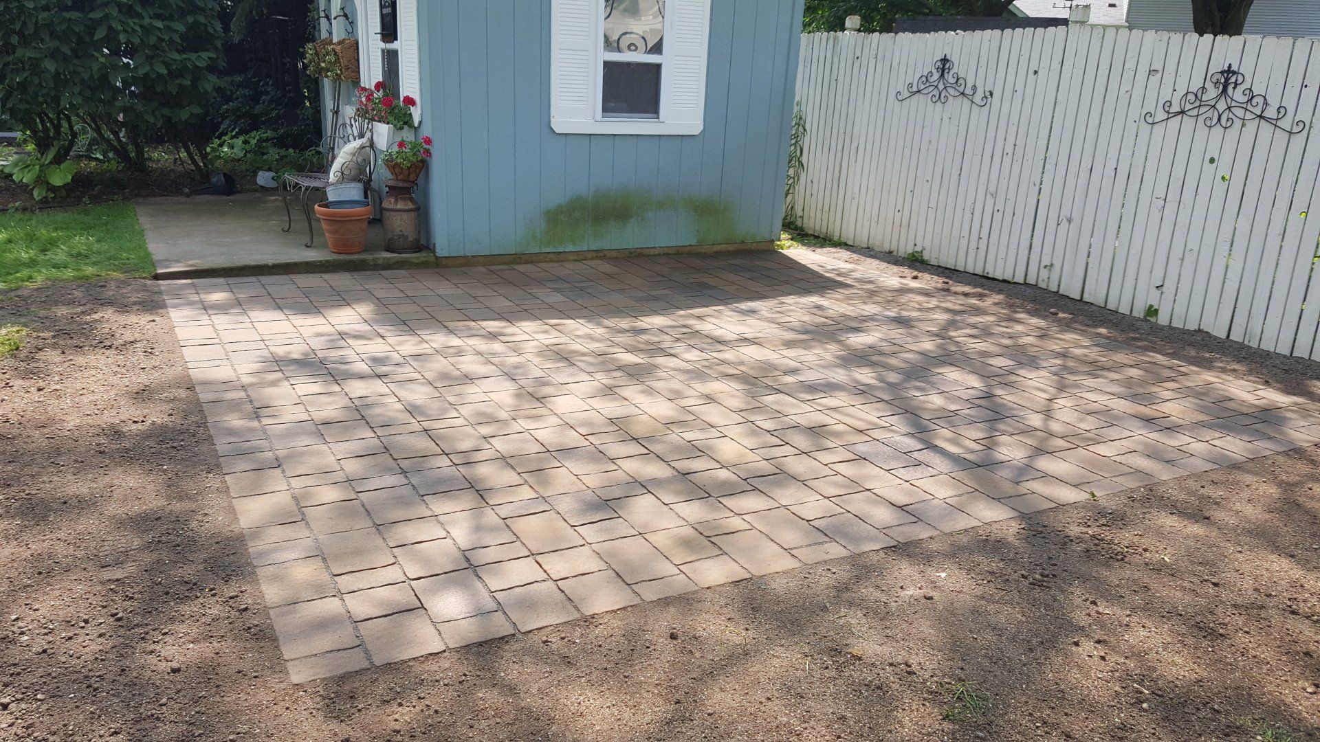 A square patio made of light-colored, perforated pavers sits in a yard next to a blue shed and a white privacy fence.