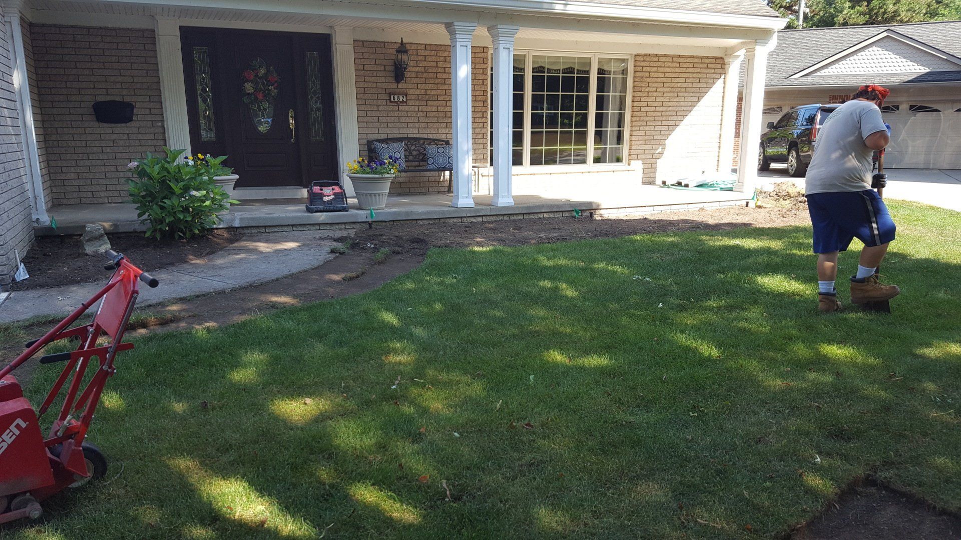A person in a gray shirt and blue shorts using a sod cutter to remove grass in front of a brick suburban house.