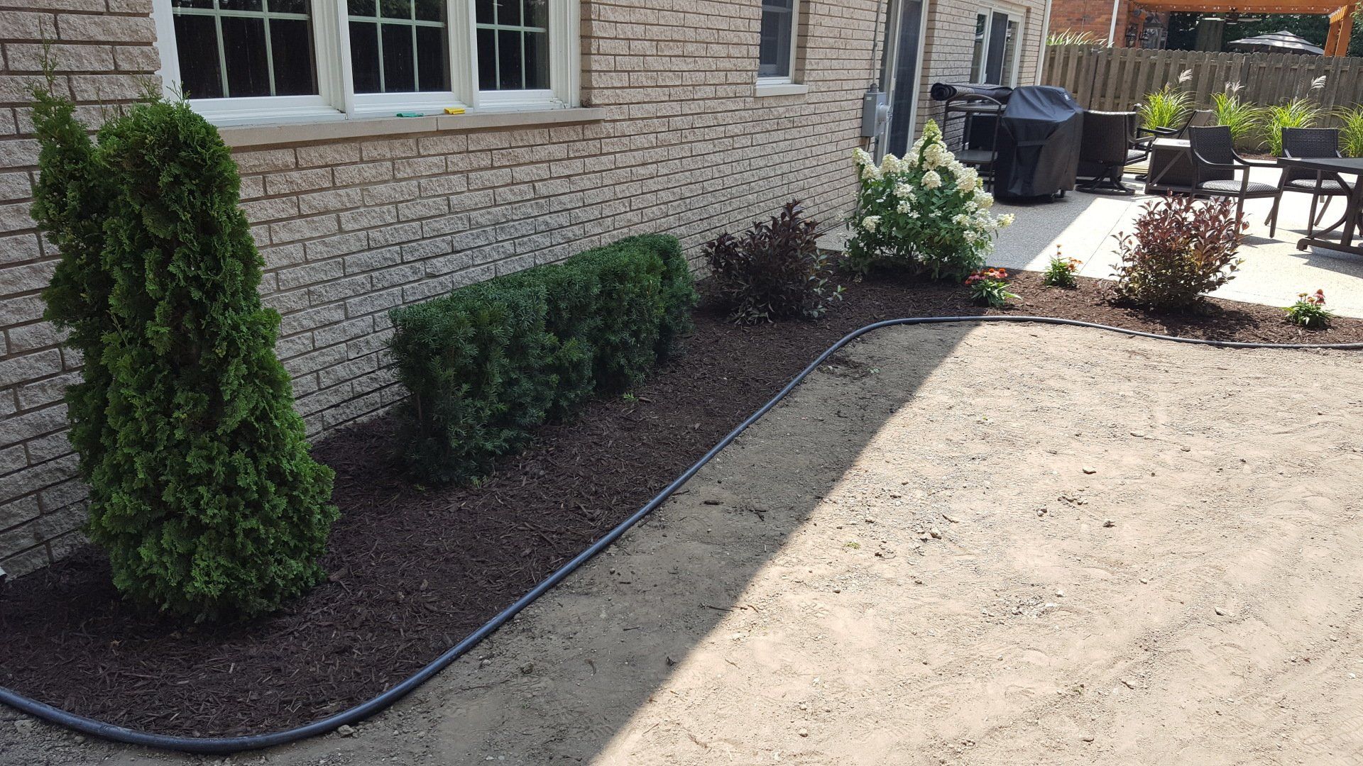 A landscaped garden bed with mulch and shrubs runs along a brick house wall next to a gravel area.