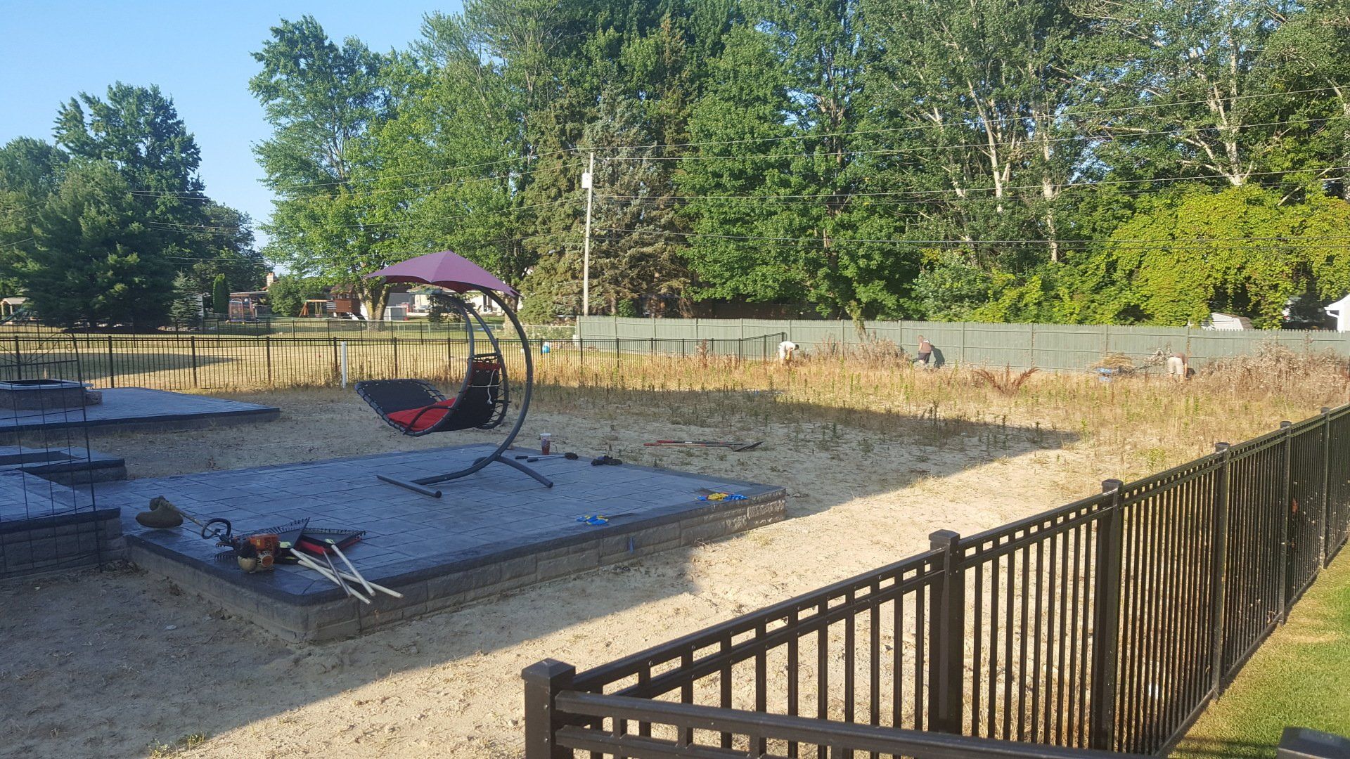 A backyard with a gravel area, a metal swing chair, and a sandy patch, enclosed by a dark fence with trees in the background.
