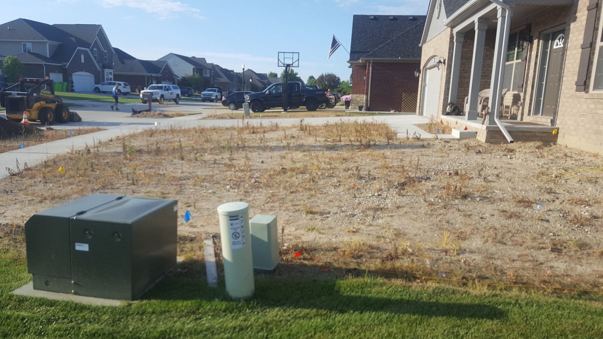 A residential front yard undergoing landscaping with exposed dirt, utility boxes in the foreground, and suburban homes.