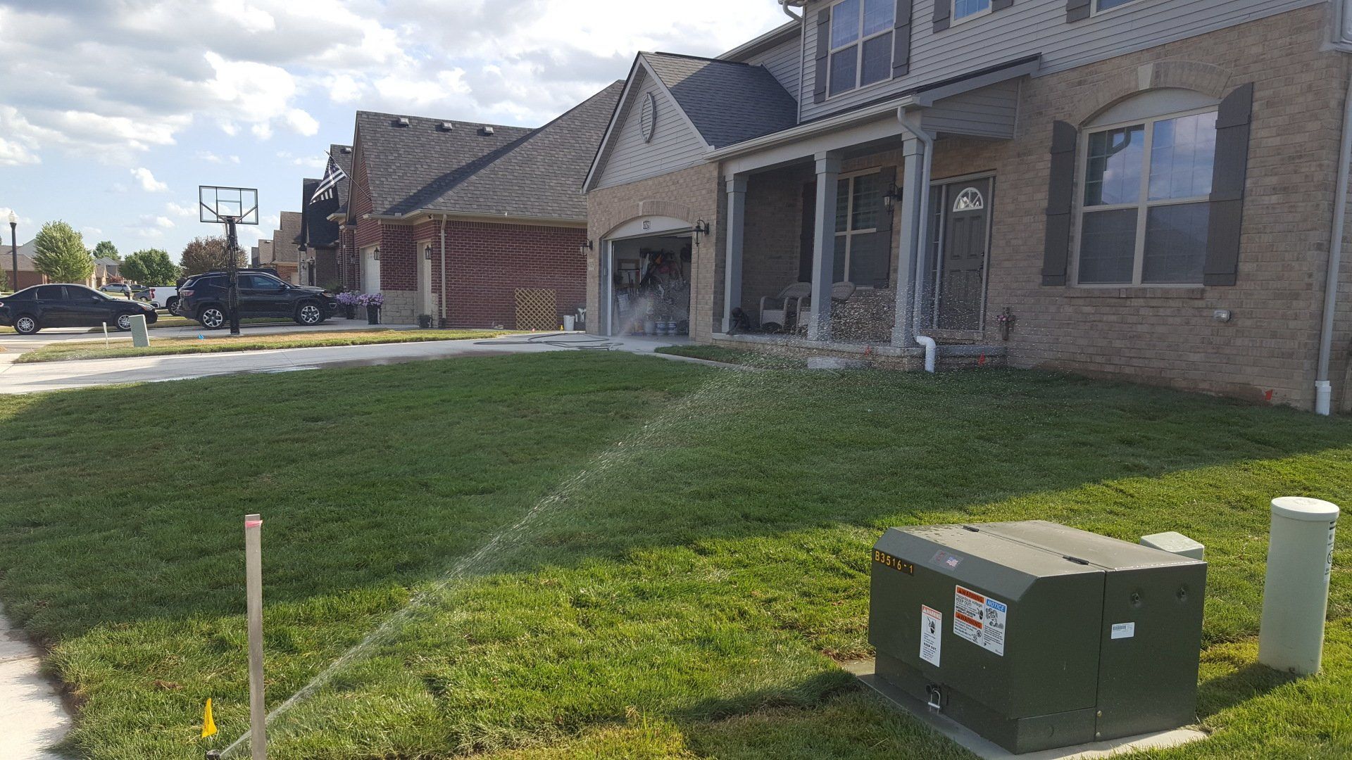 A lawn sprinkler sprays water across the green front yard of a suburban brick house on a sunny day.