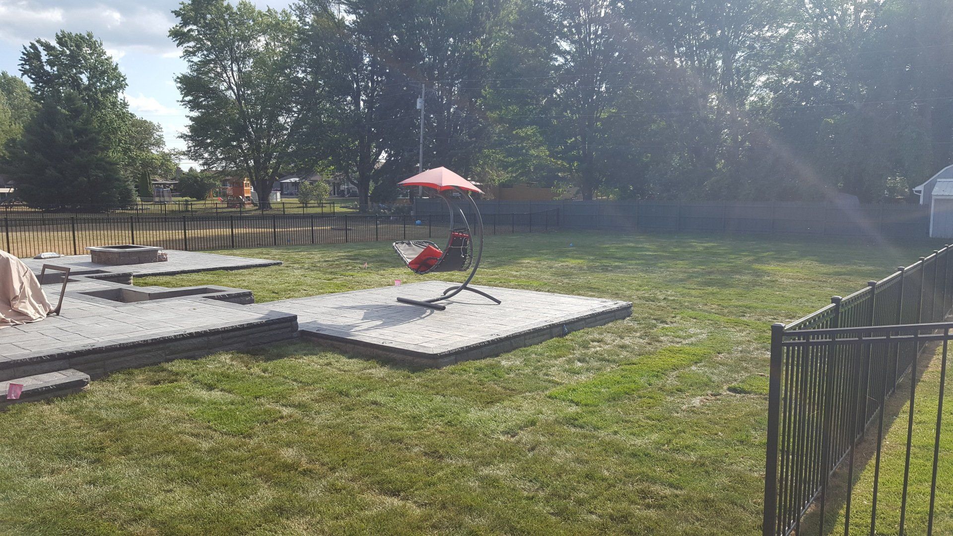 A backyard with a stone patio, a hanging chair, a fire pit, and a black fence surrounded by grass and trees.