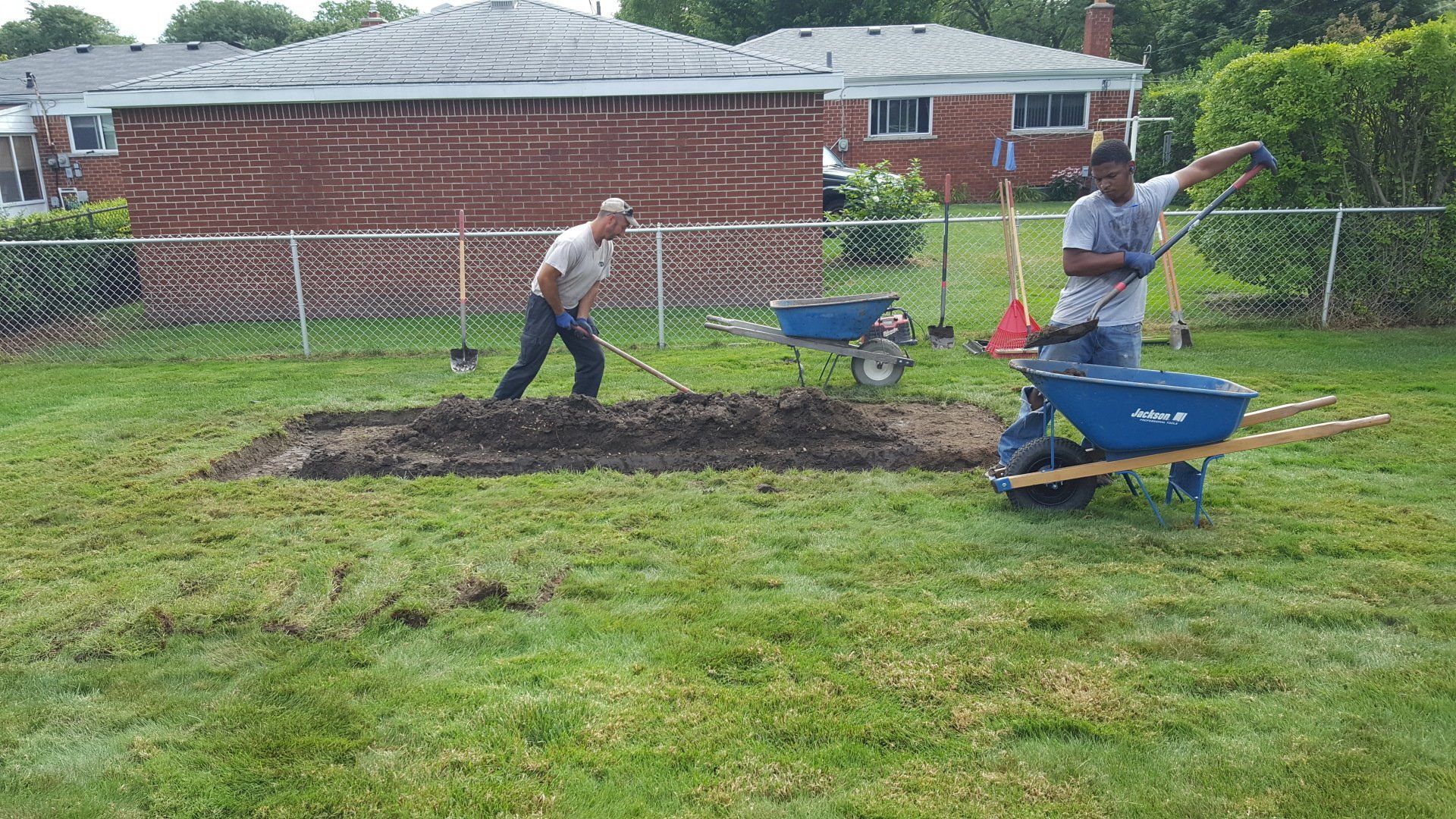 Two people work in a grassy yard, digging soil into a blue wheelbarrow near a chain-link fence and residential homes.