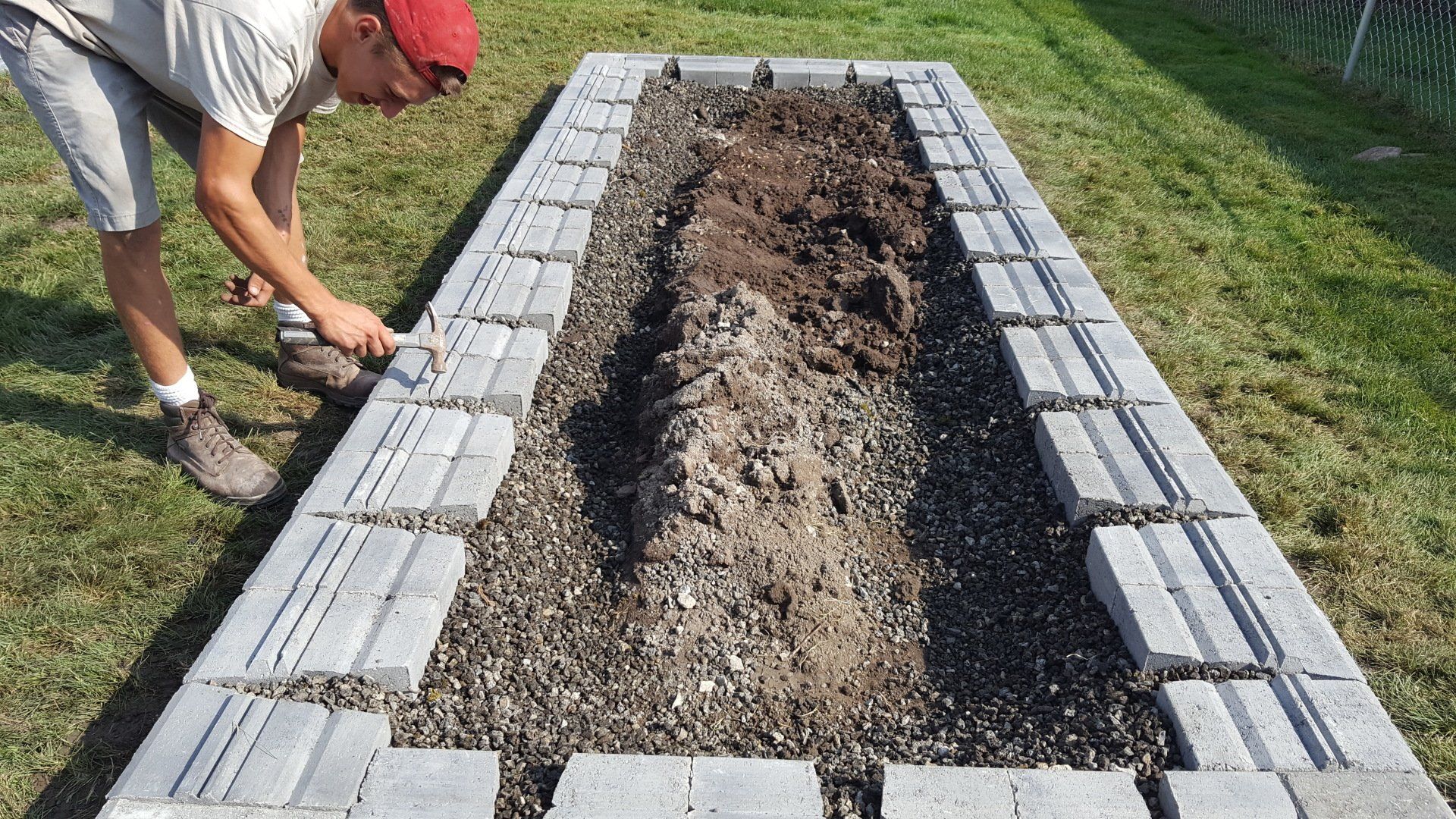 A person in a red cap aligns gray concrete blocks to build a rectangular raised garden bed in a grass yard.
