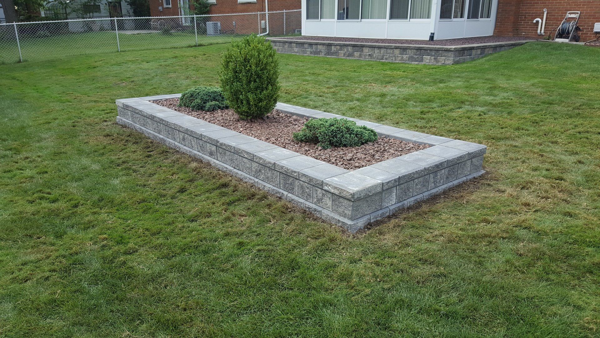 A rectangular, stone-block raised garden bed in a grassy backyard, containing gravel, one upright shrub, and two bushes.